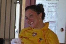 A woman is standing, smiling with her hand on her hip. She is holding a towel and wearing a bright yellow shirt.