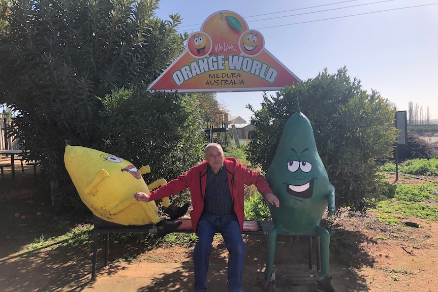 Mario Mammone sit on a park bench between large lemon and avocado characters
