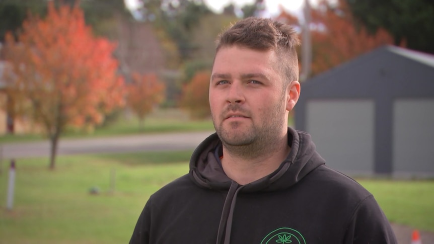 Matthew wears a black hoodie and stands near a tree with red autumn leaves.