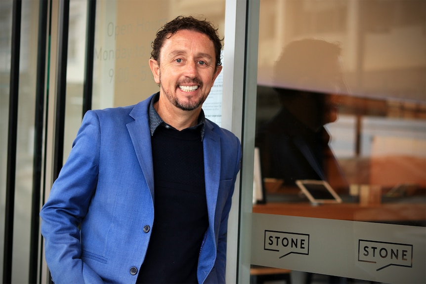 A smiling young man with a goatee wears a blue blazer and black shirt while leaning against a glass door.