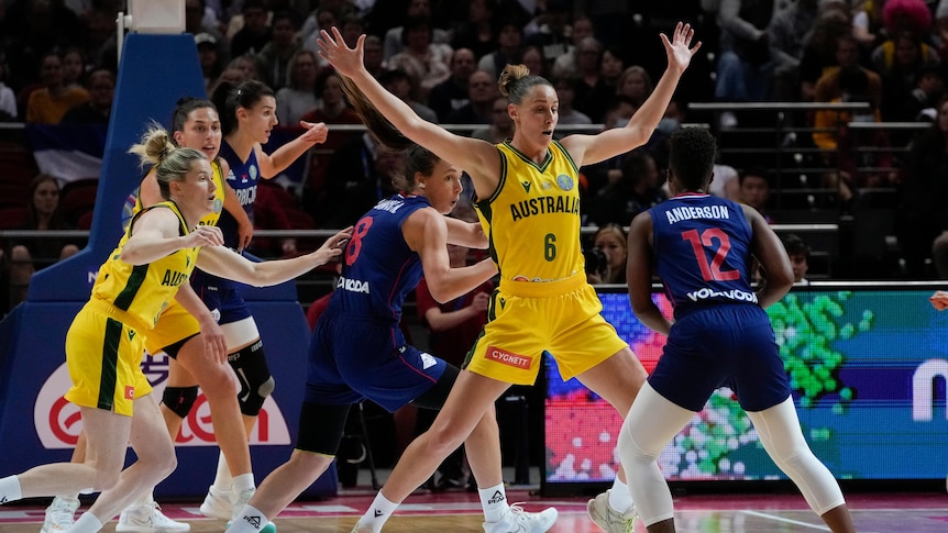 An Opals basketballer stands tall and spreads her arms wide as a Serbian player looks up ready to take a shot. 