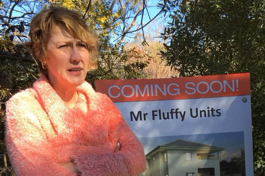 A  woman with short blonde hair stands in front of a sign that reads "Coming soon: Mr Fluffy Units".