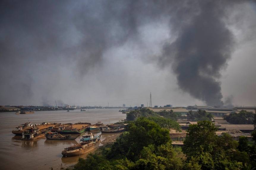 You look over a riverside group of buildings, with rusty boats by the bank, with black smoke rising behind them.