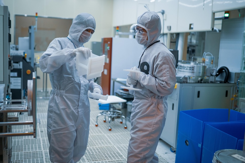 Two factory workers in white protective gear examine equipment 