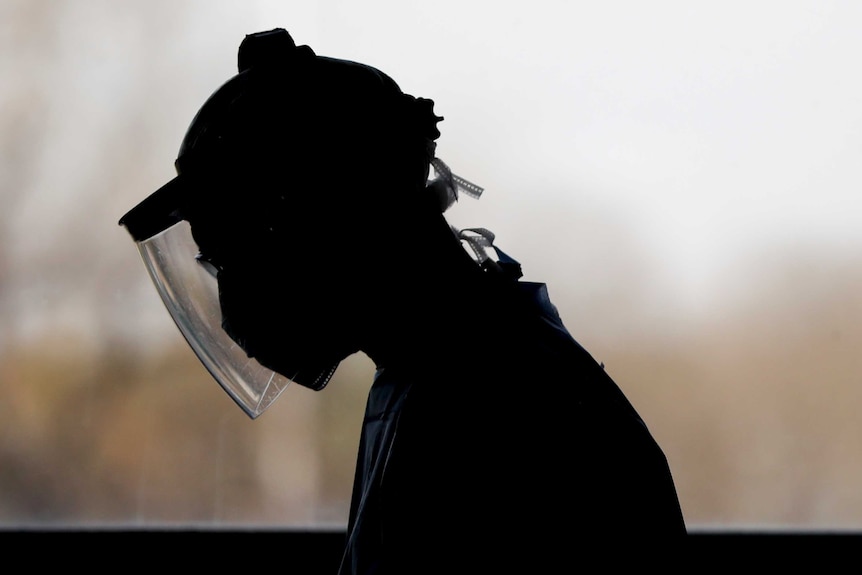 A silhouette of a doctor with head bowed wearing face shield and other PPE.