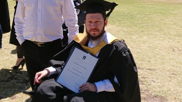 Basit sits in a wheelchair with his graduation cap and degree in hand