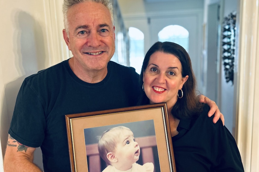A middle aged man and a woman stand together holding a picture of their baby girl.