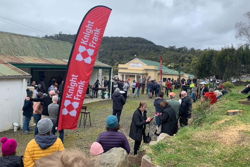 A crowd of people gather behind an old house, waiting for an auction to start.