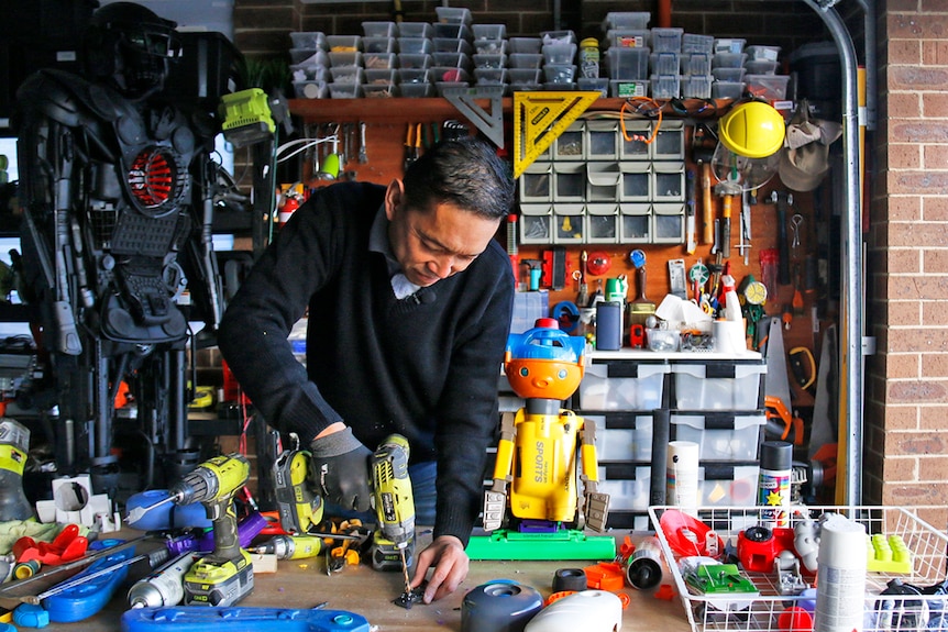 Man drilling in his garage surrounded by brightly coloured plastic parts and two robots
