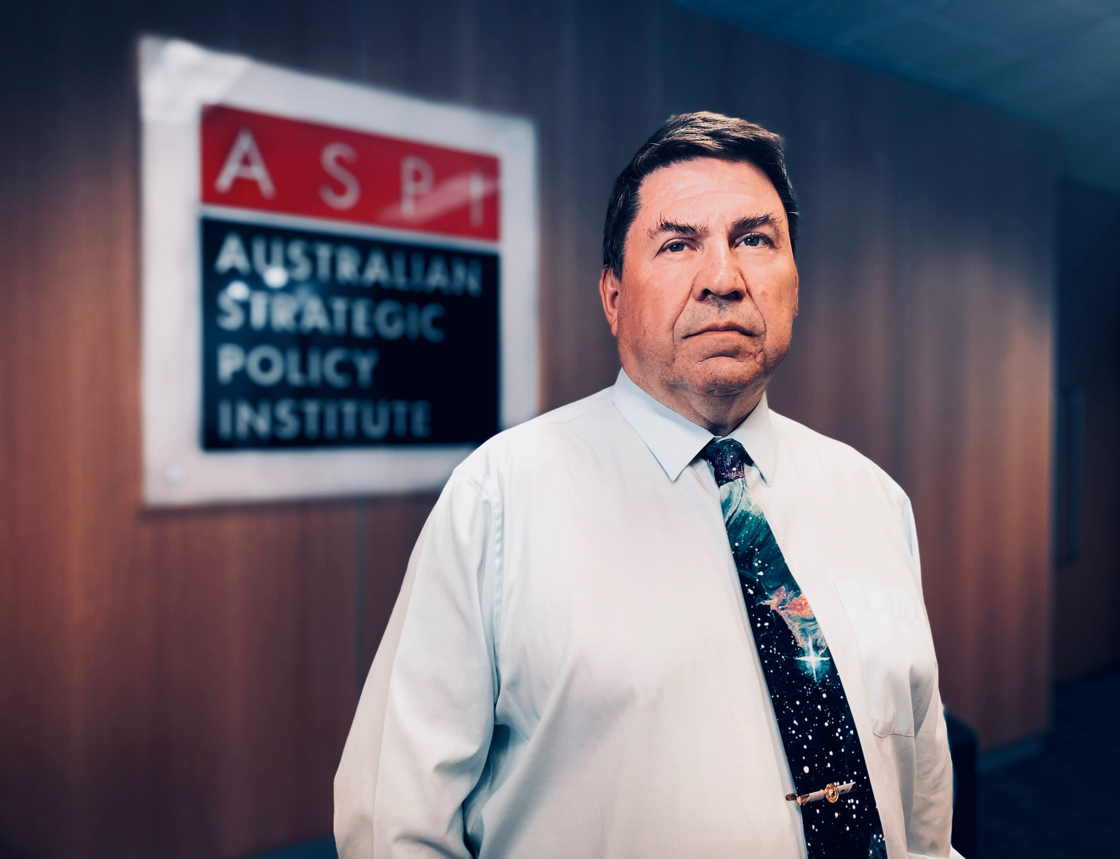 A man in a white collared shirt and tie, in the foyer of a building with the ASPI logo on the wall.