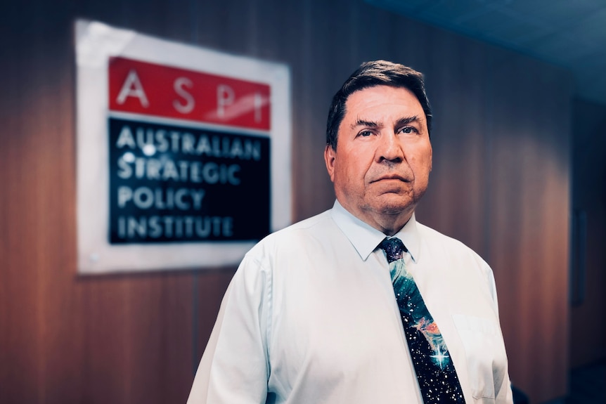 A man in a white collared shirt and tie, in the foyer of a building with the ASPI logo on the wall.