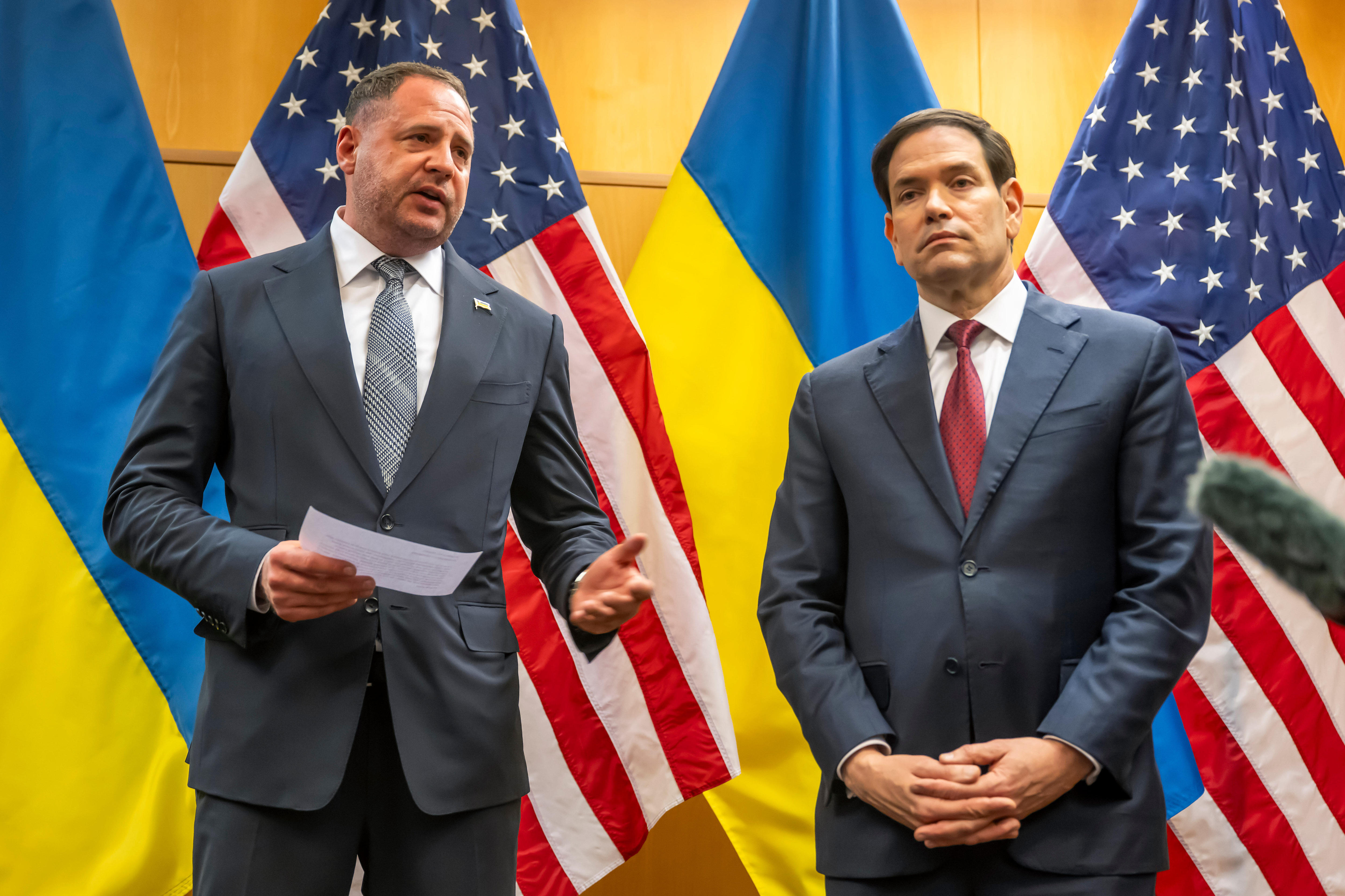 Two tall men in suits stand in front of a row of Ukrainian and American flags.
