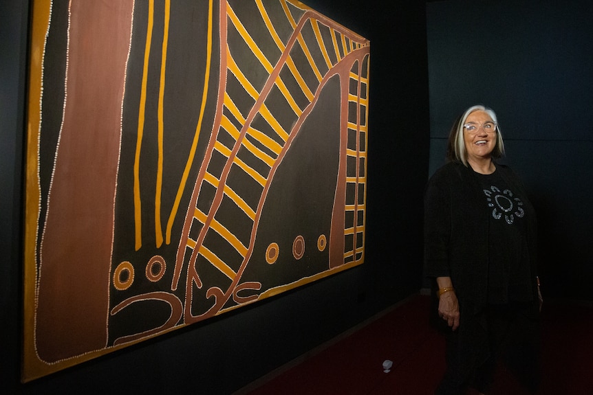 A woman with a bob stands in front of an indigenous painting.