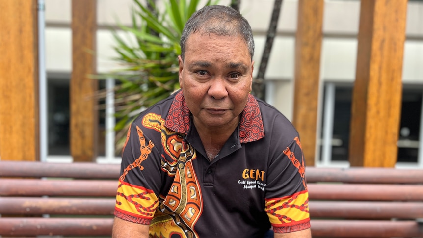 An Indigenous man with short, thinning hair, wearing a branded polo shirt, sits on a bench.