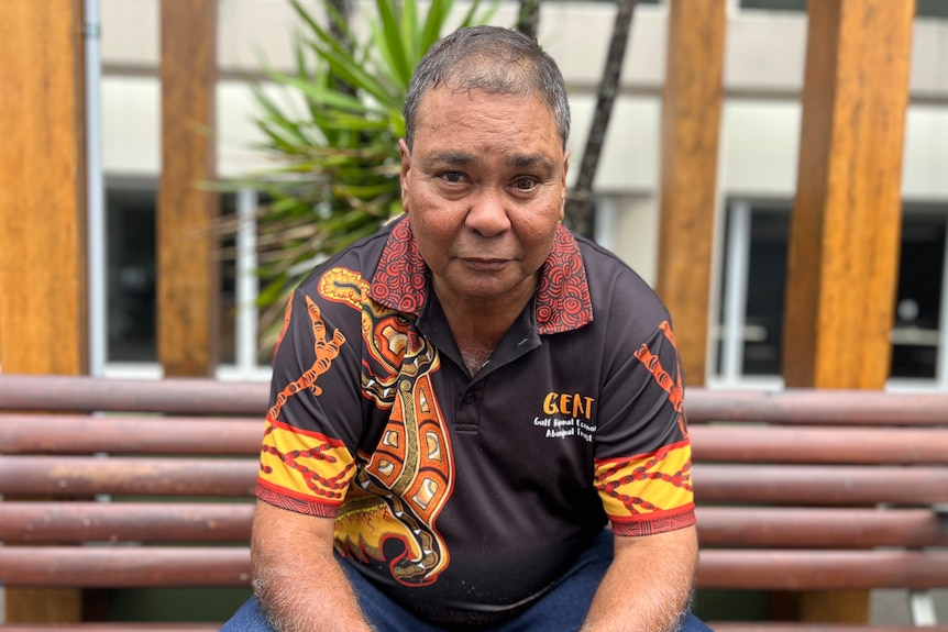 An Indigenous man with short, thinning hair, wearing a branded polo shirt, sits on a bench.