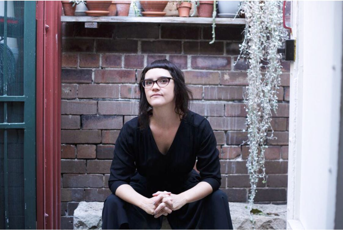 Ms Gala Vanting seated on a bench outdoors, surrounded by potplants, looking into the camera