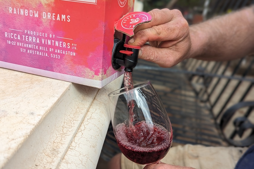 A fair-skinned hand presses a red plastic release valve to pour red wine out of a rainbow cask into a wine glass. 