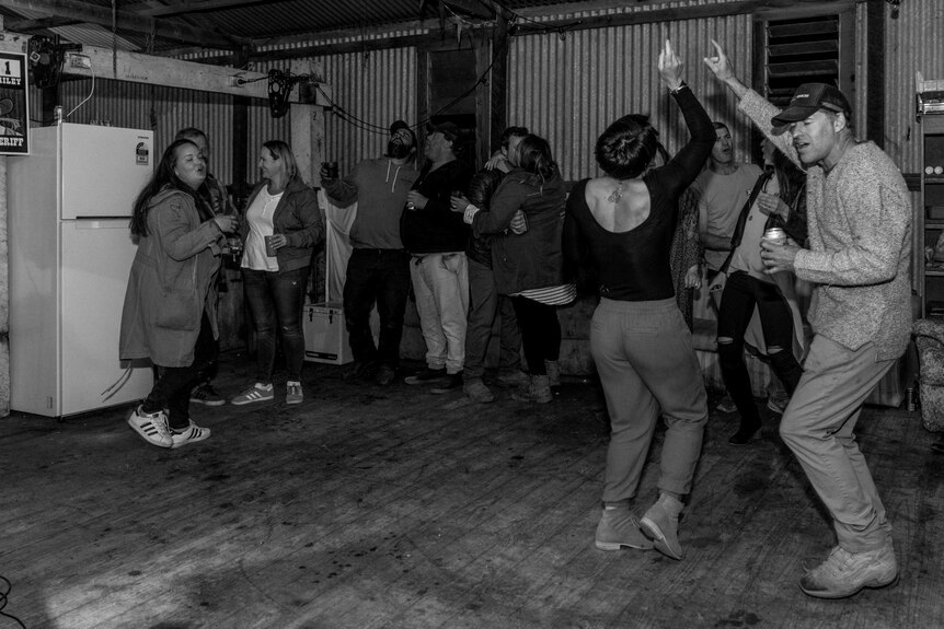 A group of people dancing and partying in a shed with a white fridge in the background