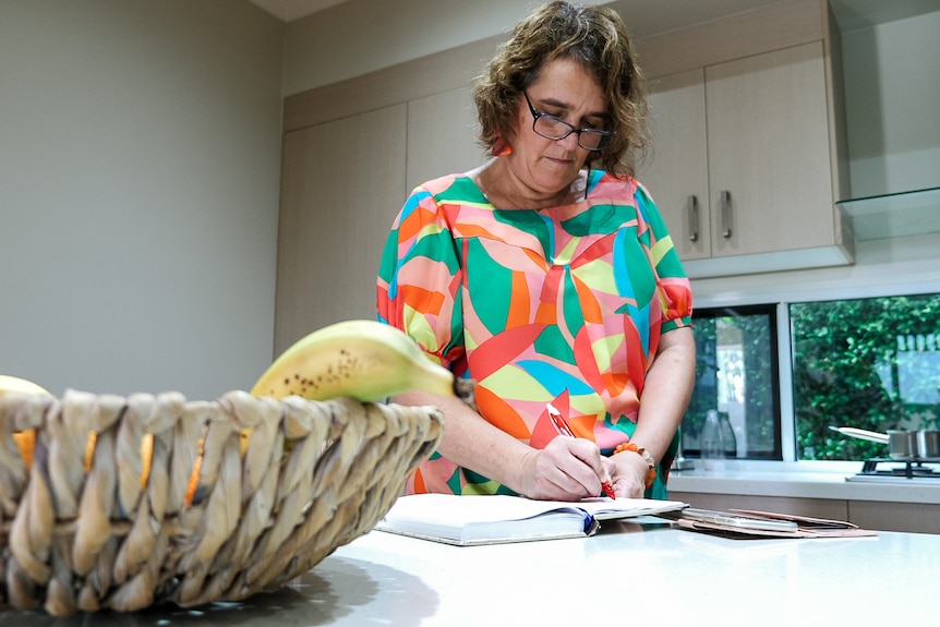 A bespectacled, middle-aged woman stands at her kitchen counter, writing in a diary.