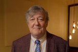 An older man with grey hair siles at the camera wearing a brown suit and a blue patterened tie. He stands in a dimly lit room.