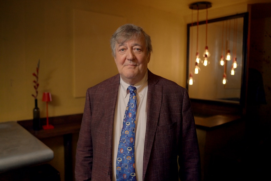 An older man with grey hair siles at the camera wearing a brown suit and a blue patterened tie. He stands in a dimly lit room.