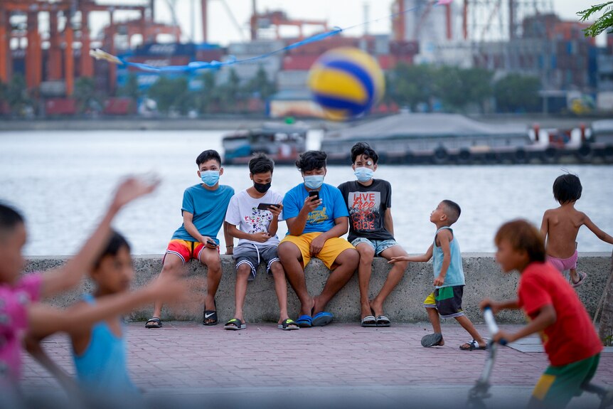 Four teen boys sit on a peer while children throw a ball in front of them 