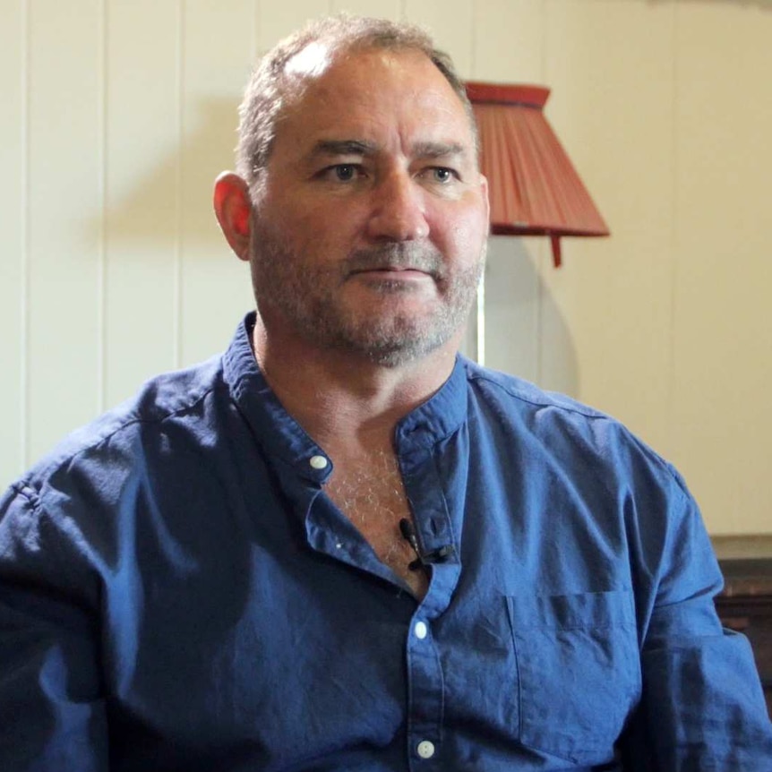 A man who looks to be in his mid-40s with grey facial hair sits in a chair in an older house.