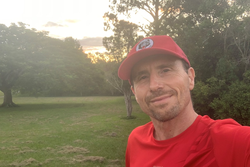 A 50-something man in red shirt and red cap stands in a park in front of a sunset