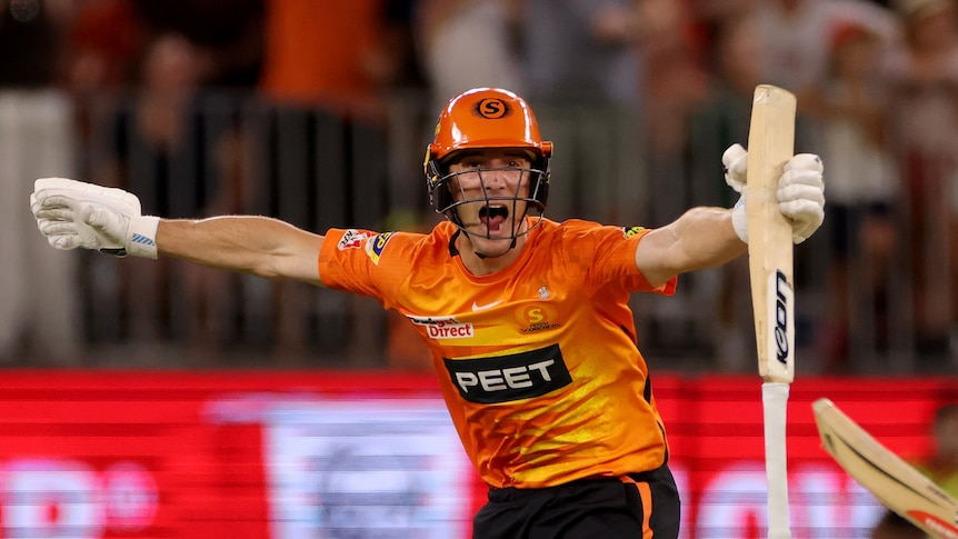A man in an orange shirt holding a cricket bat celebrates with arms outstretched