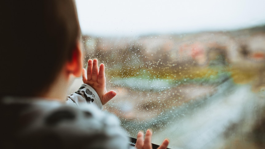 A small child puts its hands on the window inside a house and looks outside. 