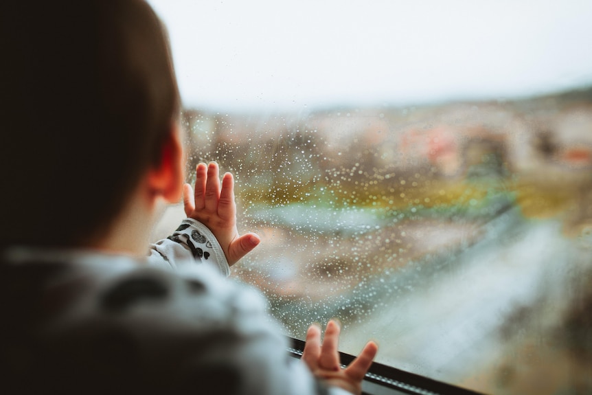 A small child puts its hands on the window inside a house and looks outside. 