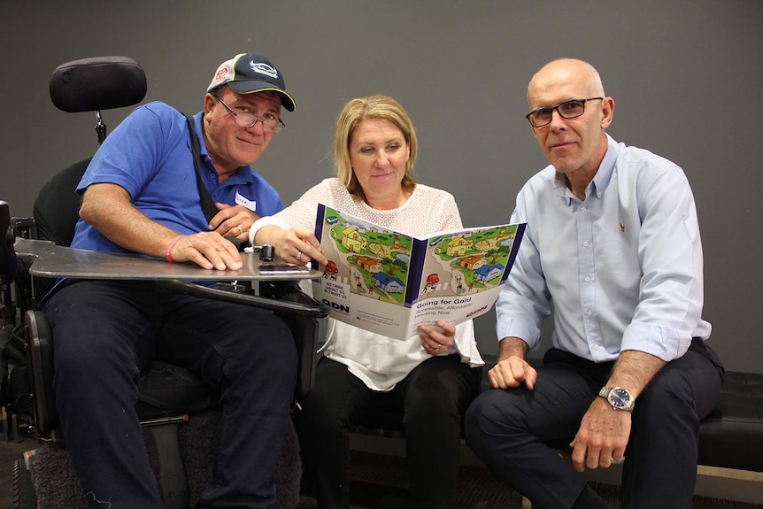 Three people sit on a couch reading a booklet on accessible housing. One person is in a wheelchair.