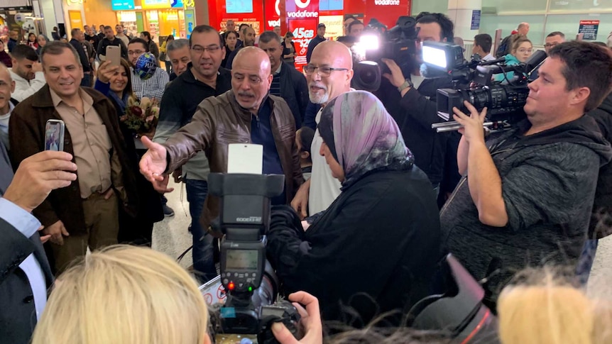 A bald man wearing glasses is surrounded by people and television crews and photographers.