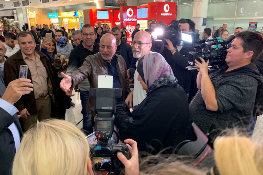A bald man wearing glasses is surrounded by people and television crews and photographers.
