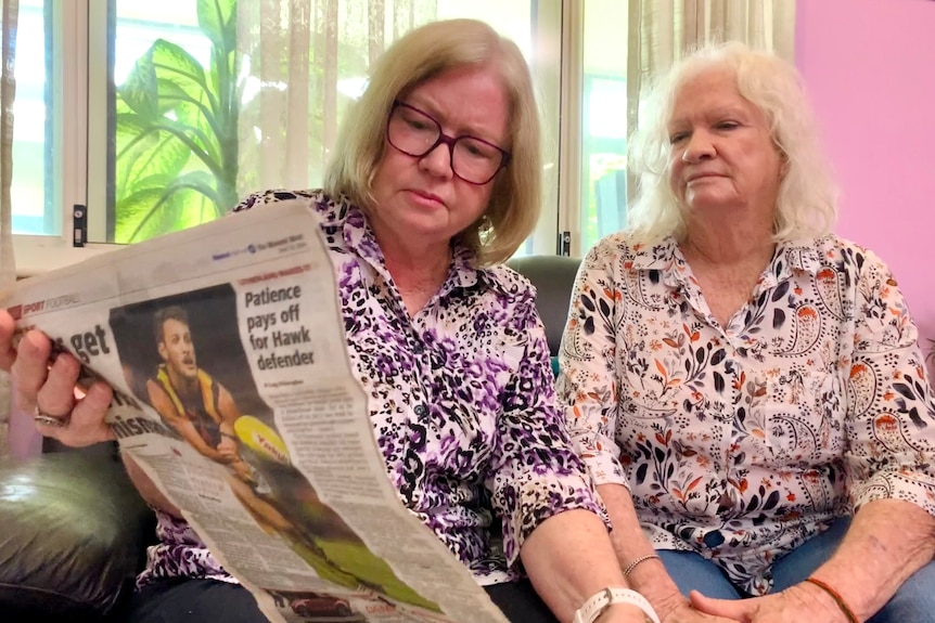 Two women study a old newspaper article in a sunny living room