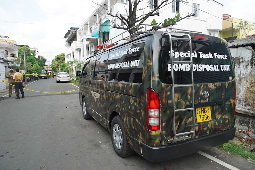 A camouflage van is parked in front of a white house, with police in the background with yellow crime scene tape.