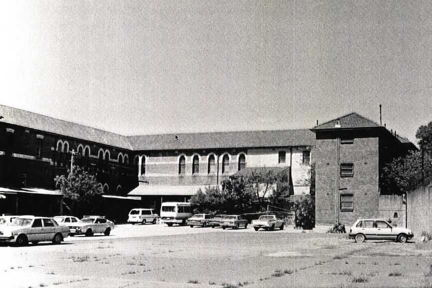 Grainy, black and white photo of a large brick building with arched windows, and several cars parked out the front of it.
