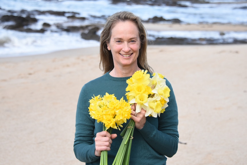 A woman stands on a beach holding multiple bunches of daffodils.
