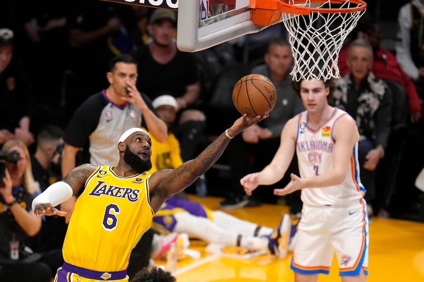 Basketballer LeBron James reaches out for a lay-up shot to the basket as defender Josh Giddey watches in the background.