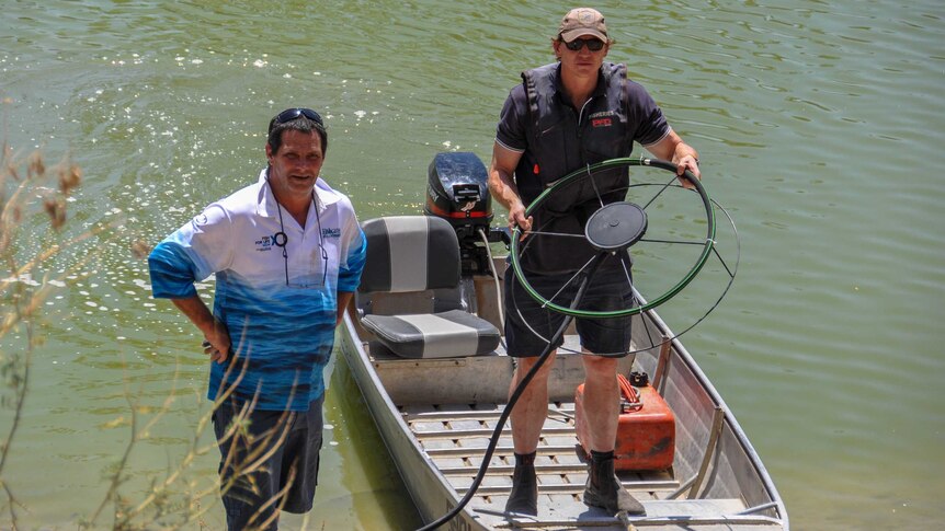 Two men stand on the bank of a waterway. One is in a boat holding up a fan-like object.