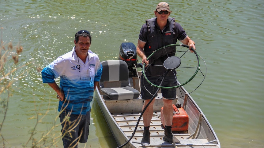 Two men stand on the bank of a waterway. One is in a boat holding up a fan-like object.