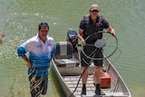 Two men stand on the bank of a waterway. One is in a boat holding up a fan-like object.