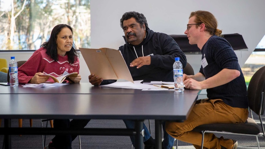 Three people sit at a desk gesticulating