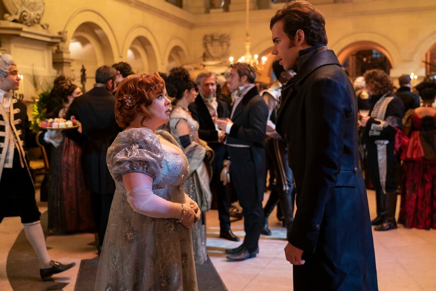 The characters of Penelope Featherington and Colin Bridgerton stand contemplating each other at a ball.