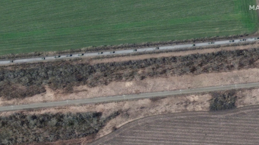 A bird's eye view of a number of a khaki vehicles moving along a road with a green field on one side and dry shrubs on the other