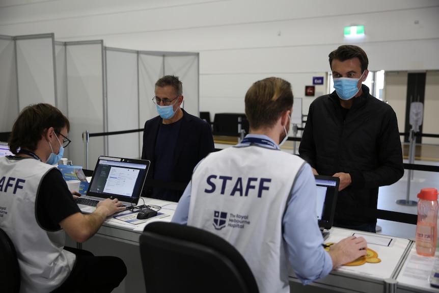 Two men stand at a desk speaking to people wearing white vests that say staff.