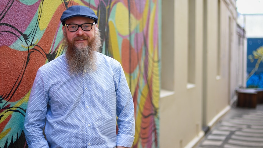A man with glasses and a beard, wearing a flat cap and a blue shirt, standing in a laneway near a mural and a historic building.
