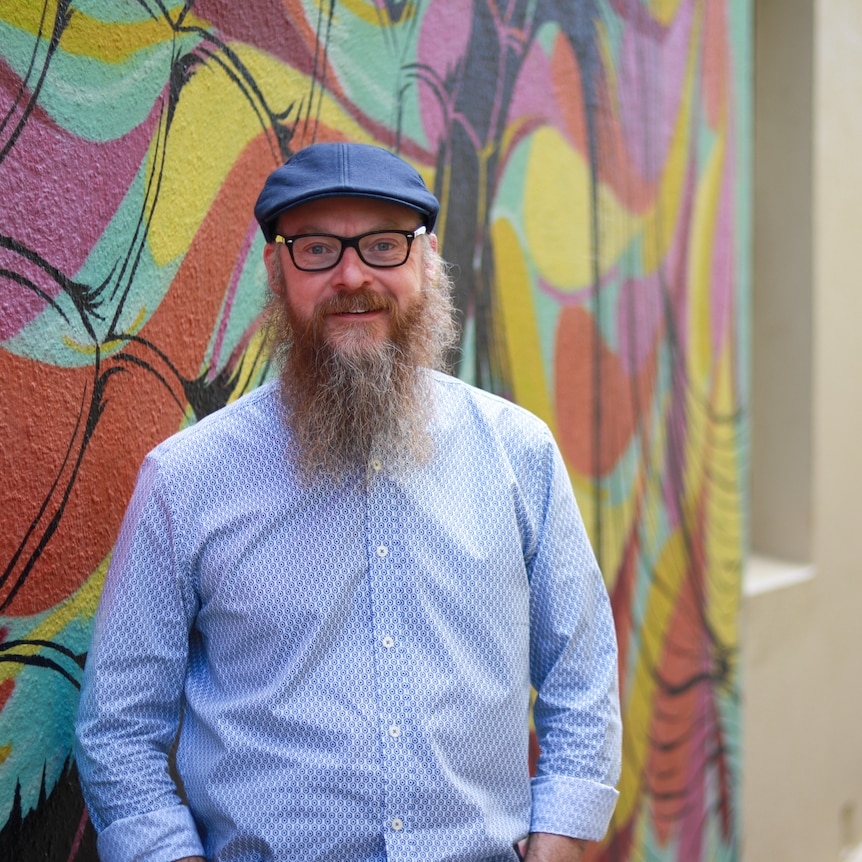 A man with glasses and a beard, wearing a flat cap and a blue shirt, standing in a laneway near a mural and a historic building.