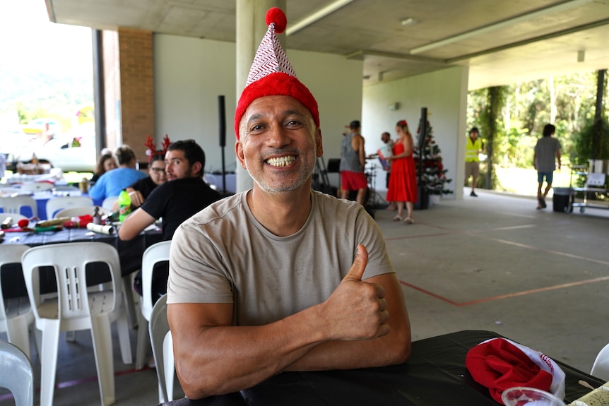 A man with Christmas hat sitting at table smiles and gives the thumbs up.