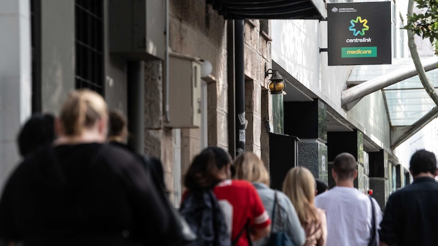 A line of people photographed from behind in front of a sign that reads Centrelink.